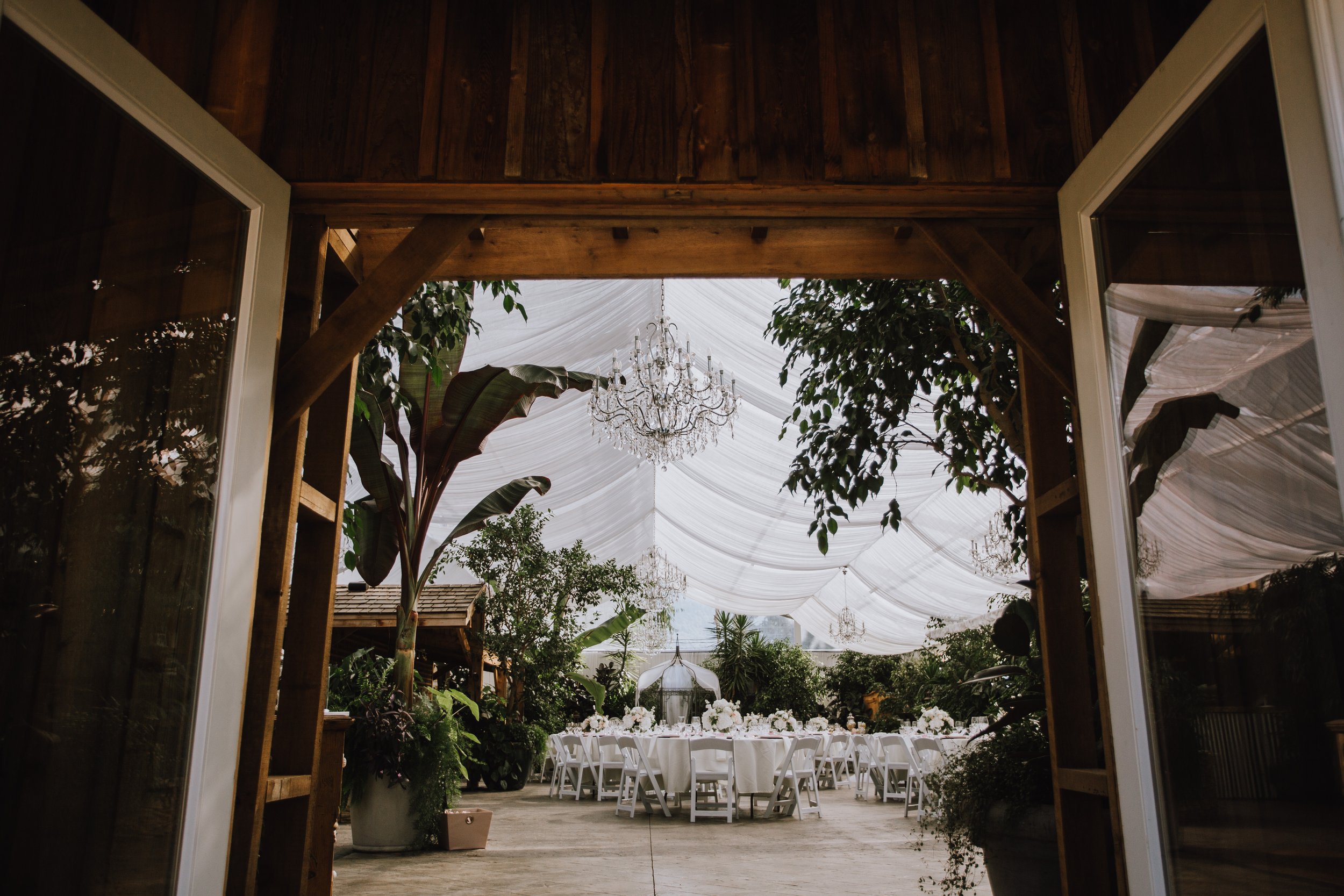 Elegant outdoor event space with white draped ceiling, chandeliers, and round tables with white tablecloths and floral centerpieces, surrounded by lush greenery.