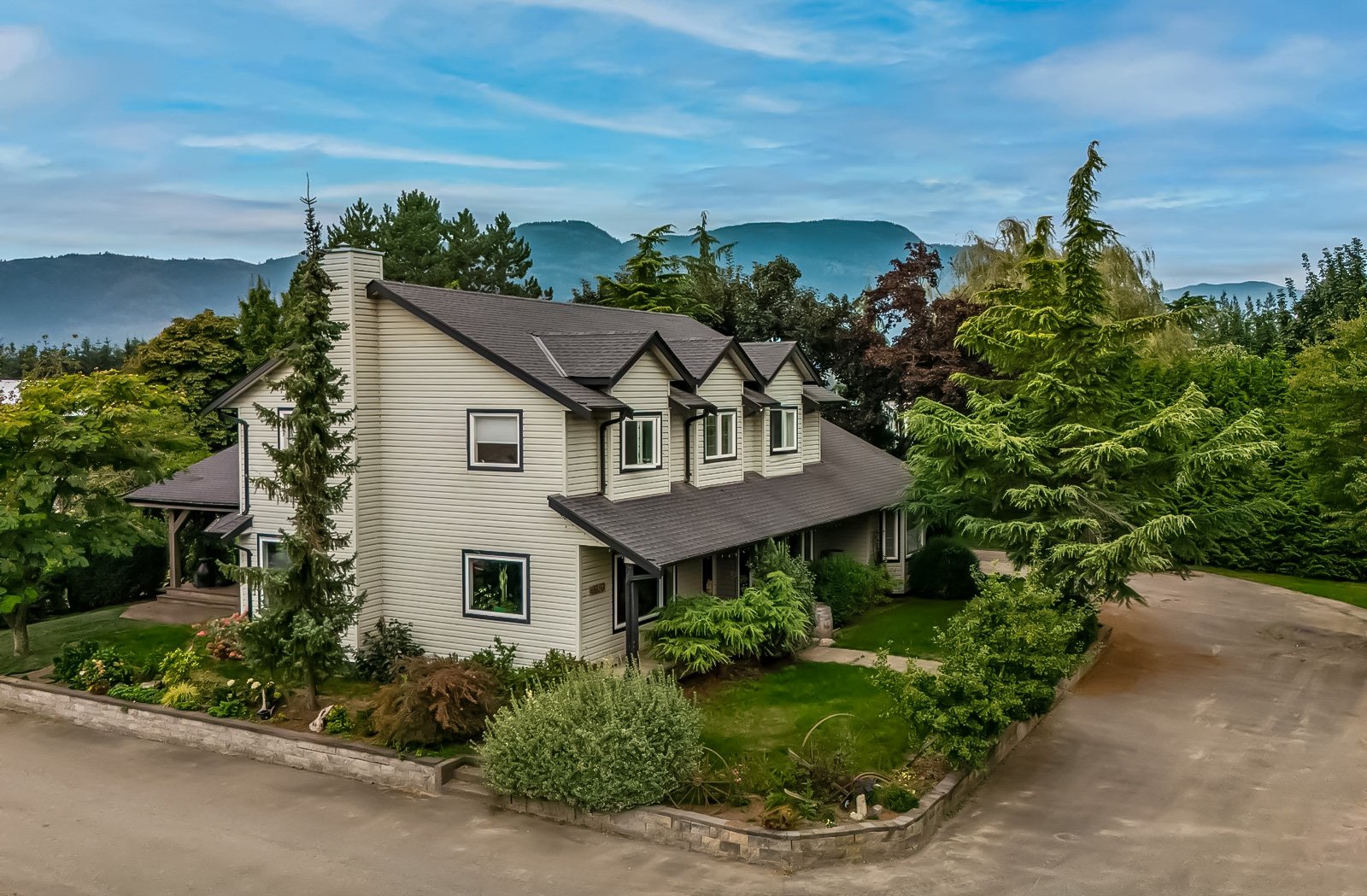 A two-story house with beige siding, dark gray roof, and multiple windows, surrounded by lush greenery and trees, with mountains in the background under a partly cloudy sky.