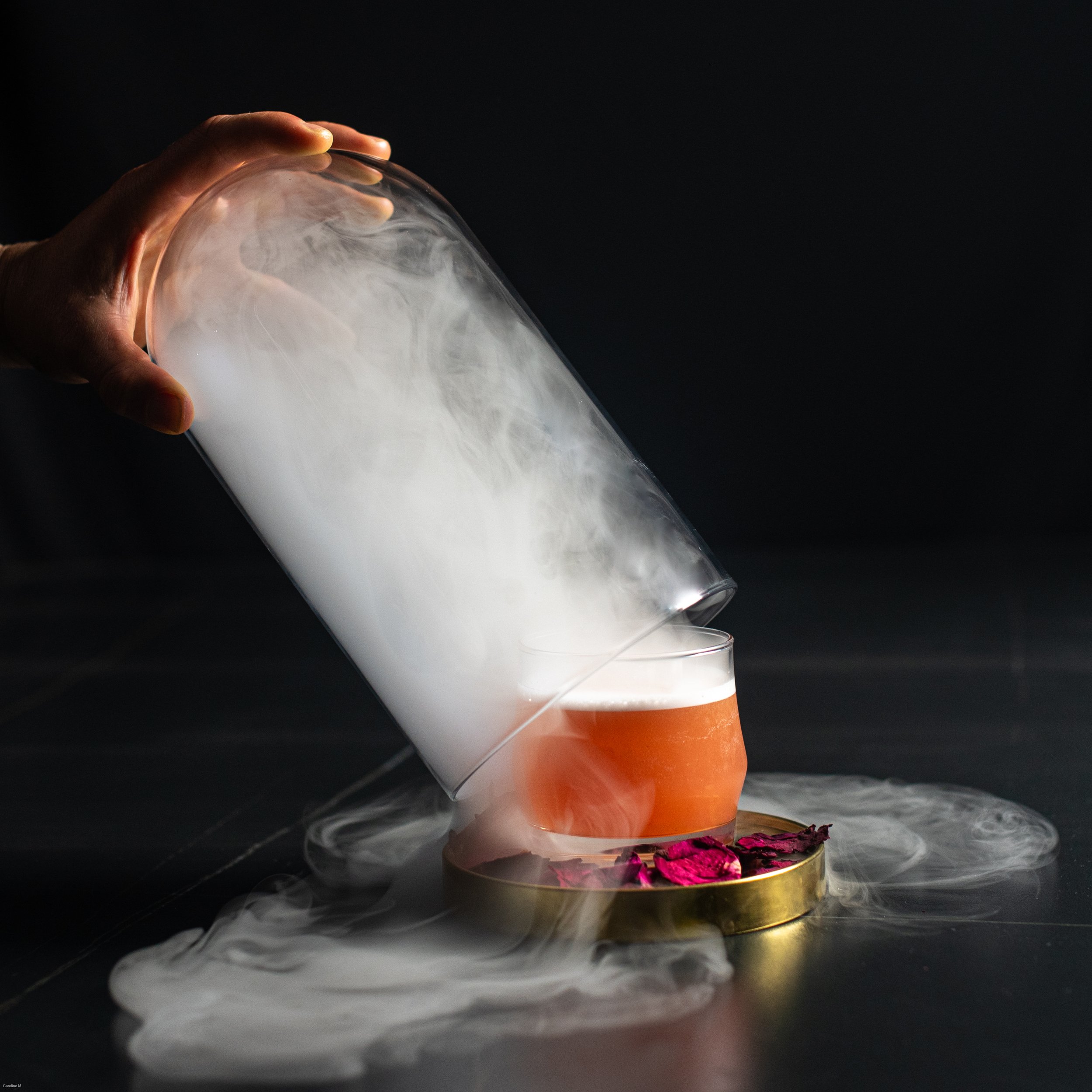Hand holding a glass container with white smoke and pouring into a glass with pink beverage, on a black surface with scattered pink flower petals