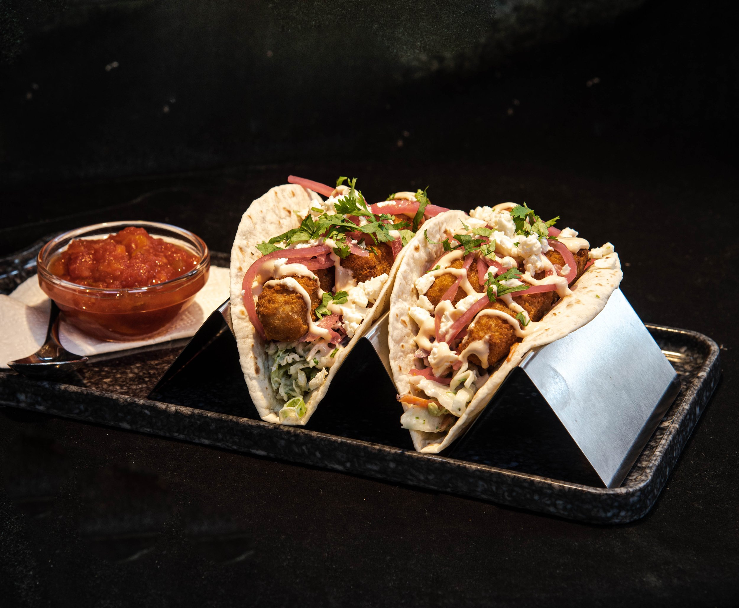 Two fish tacos in soft tortillas filled with breaded fish, shredded lettuce, red onion, cilantro, and drizzled with sauce, served with a side of salsa in a small glass bowl on a black tray.