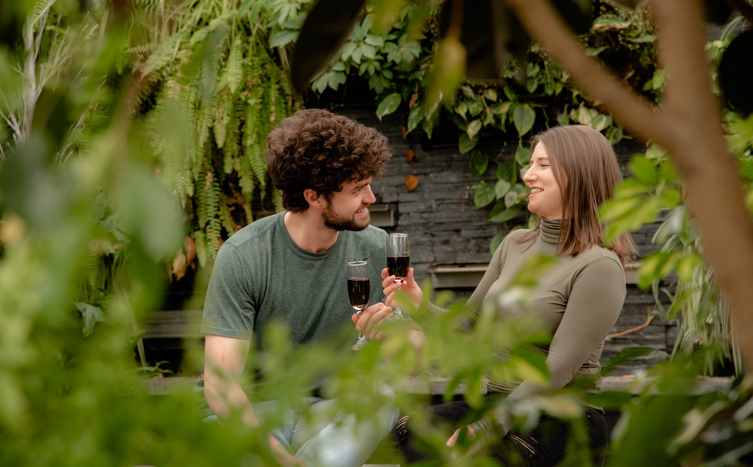A man and woman sitting outdoors surrounded by green plants, smiling and clinking glasses of red wine.