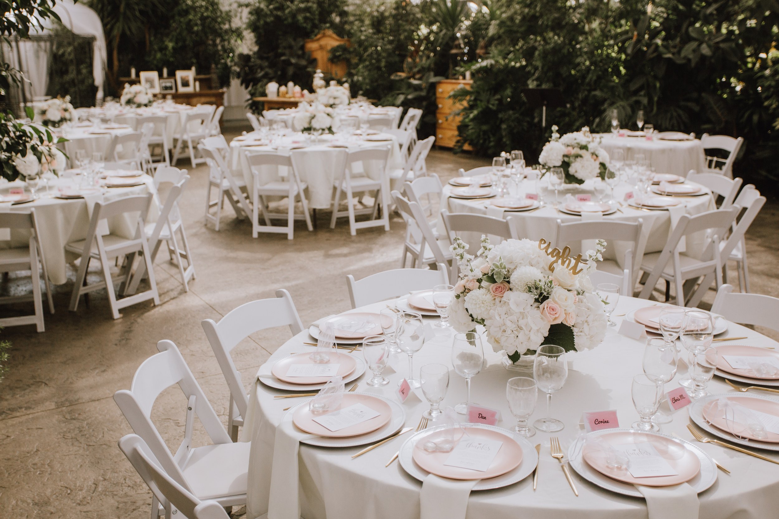 Elegant indoor wedding reception setup with round tables decorated with white tablecloths, pink plates, gold utensils, and glassware. Each table has a floral centerpiece with white and blush flowers, and pink name cards. The background features lush greenery and framed photographs.