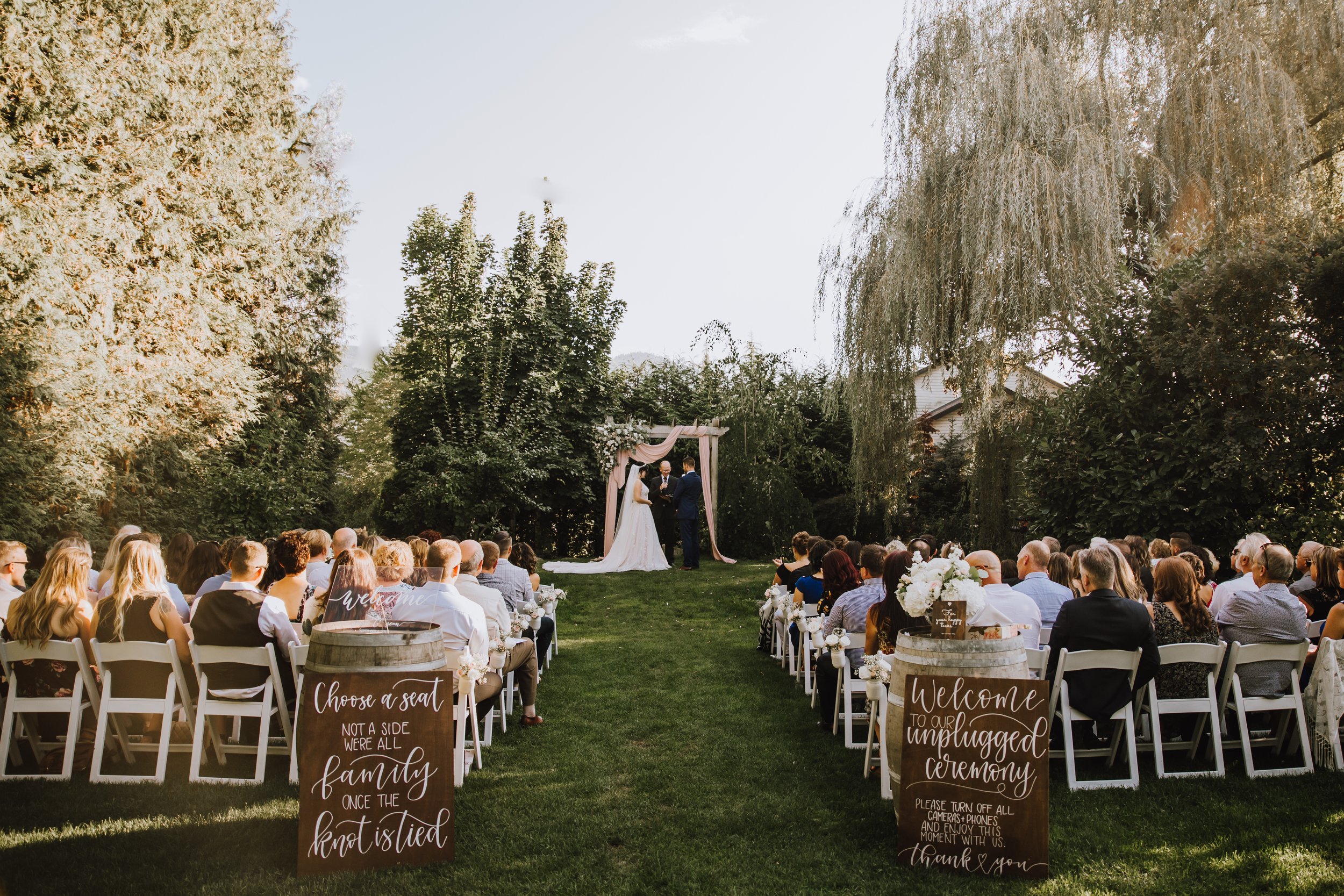 Outdoor wedding ceremony with guests seated on white chairs, two signs on wooden barrels with welcoming messages, and a couple standing under a pink draped arch.