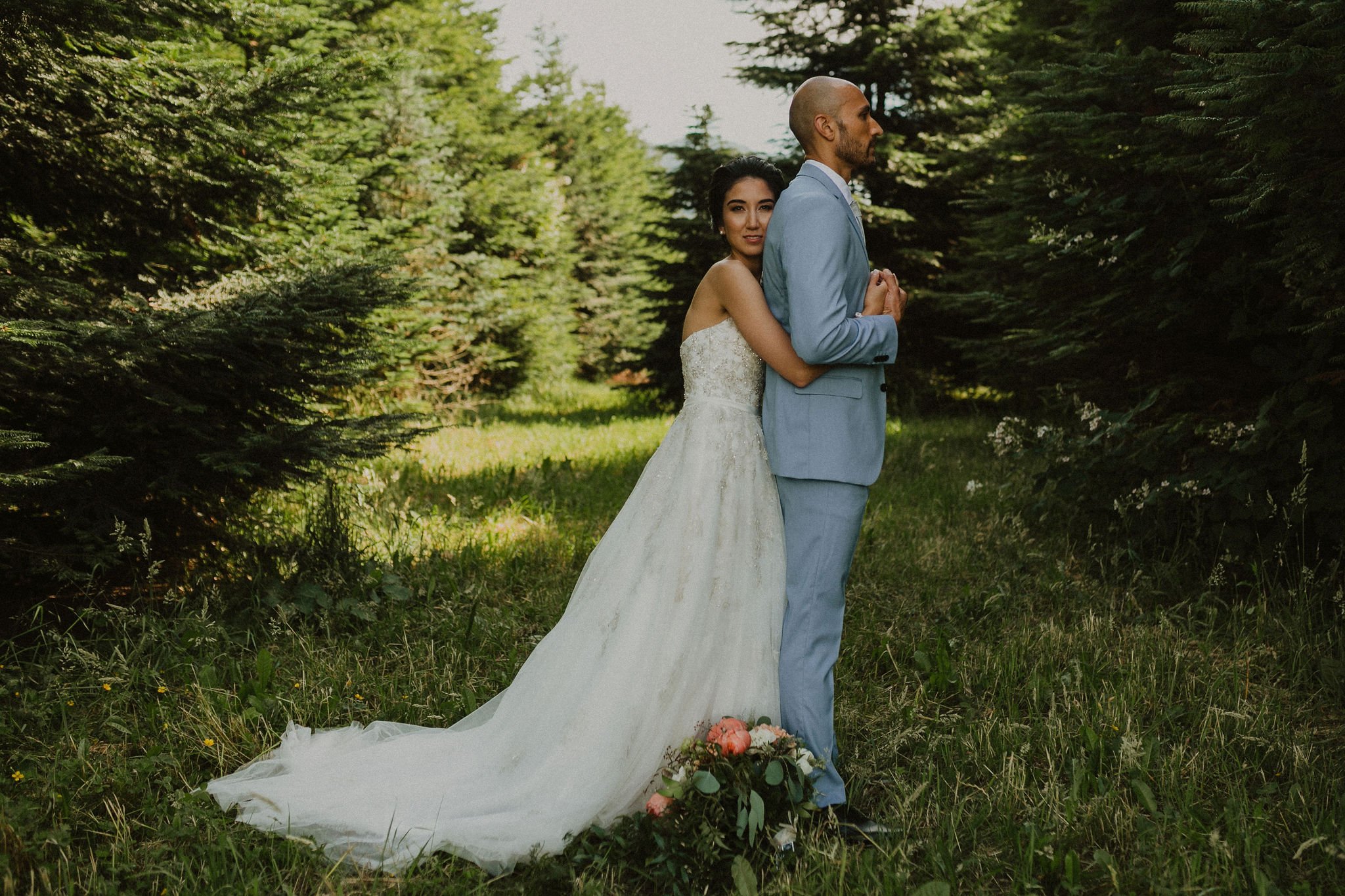 A bride in a white wedding dress hugging a groom in a light blue suit, standing outdoors on a grassy path surrounded by evergreen trees.