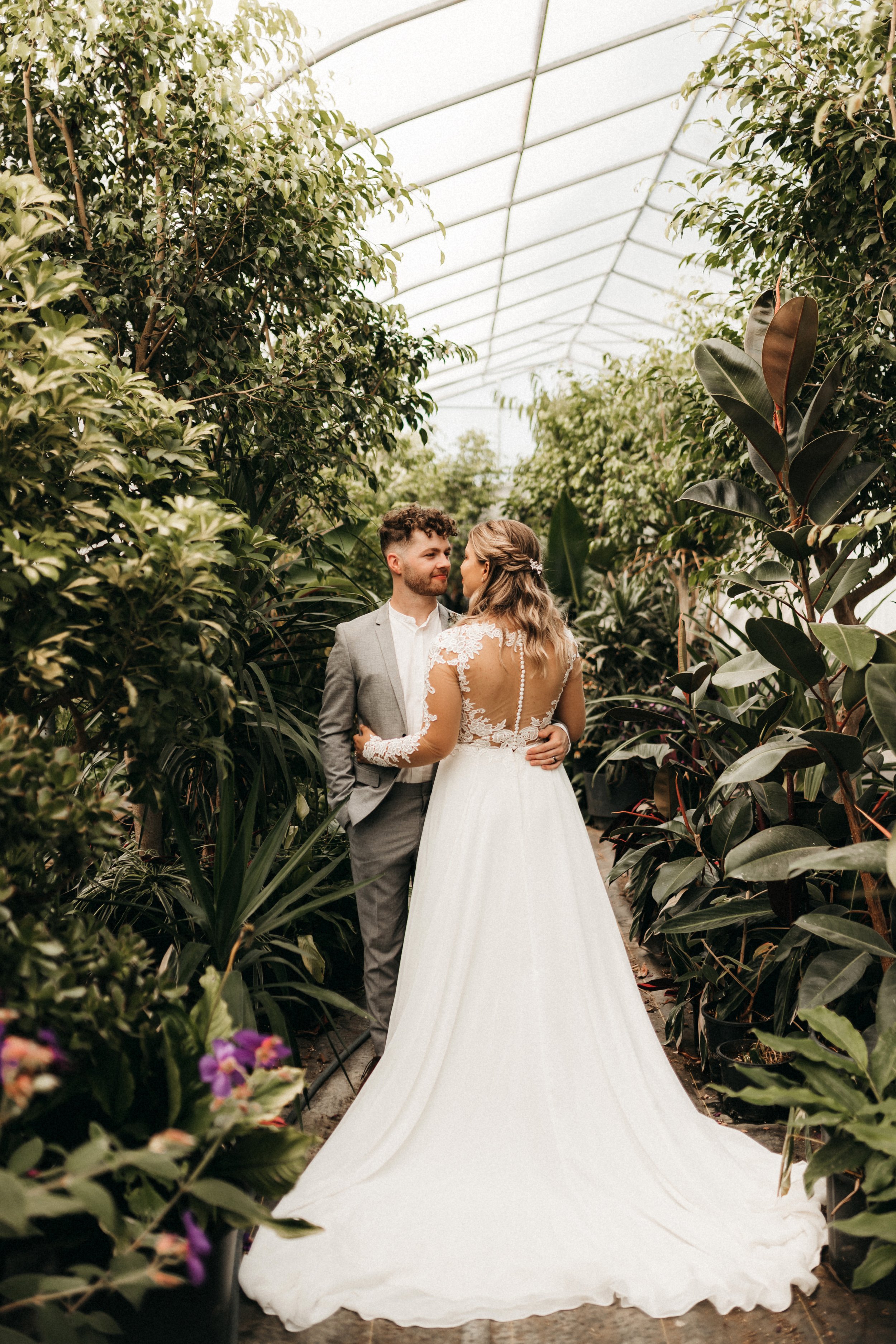 A bride and groom embracing in a glass greenhouse, surrounded by lush green plants. The bride wears a white wedding gown with lace details, and the groom is dressed in a gray suit with a white shirt.