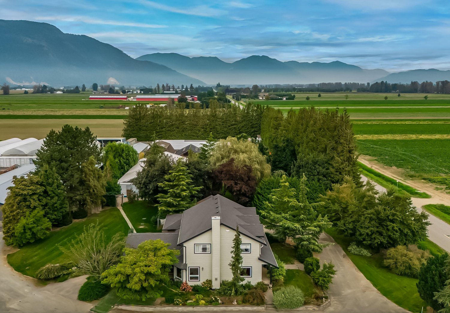 Aerial view of a house surrounded by lush green trees in a rural area with farmland and mountains in the background under a partly cloudy sky.
