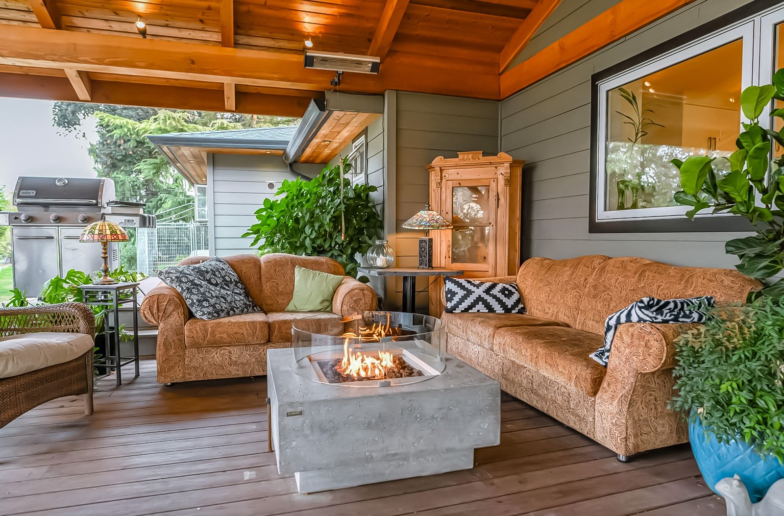 Indoor living space with wicker and upholstered sofas, a stone fire pit, plants, a wooden cabinet, and large windows showing an outdoor grill and green trees.