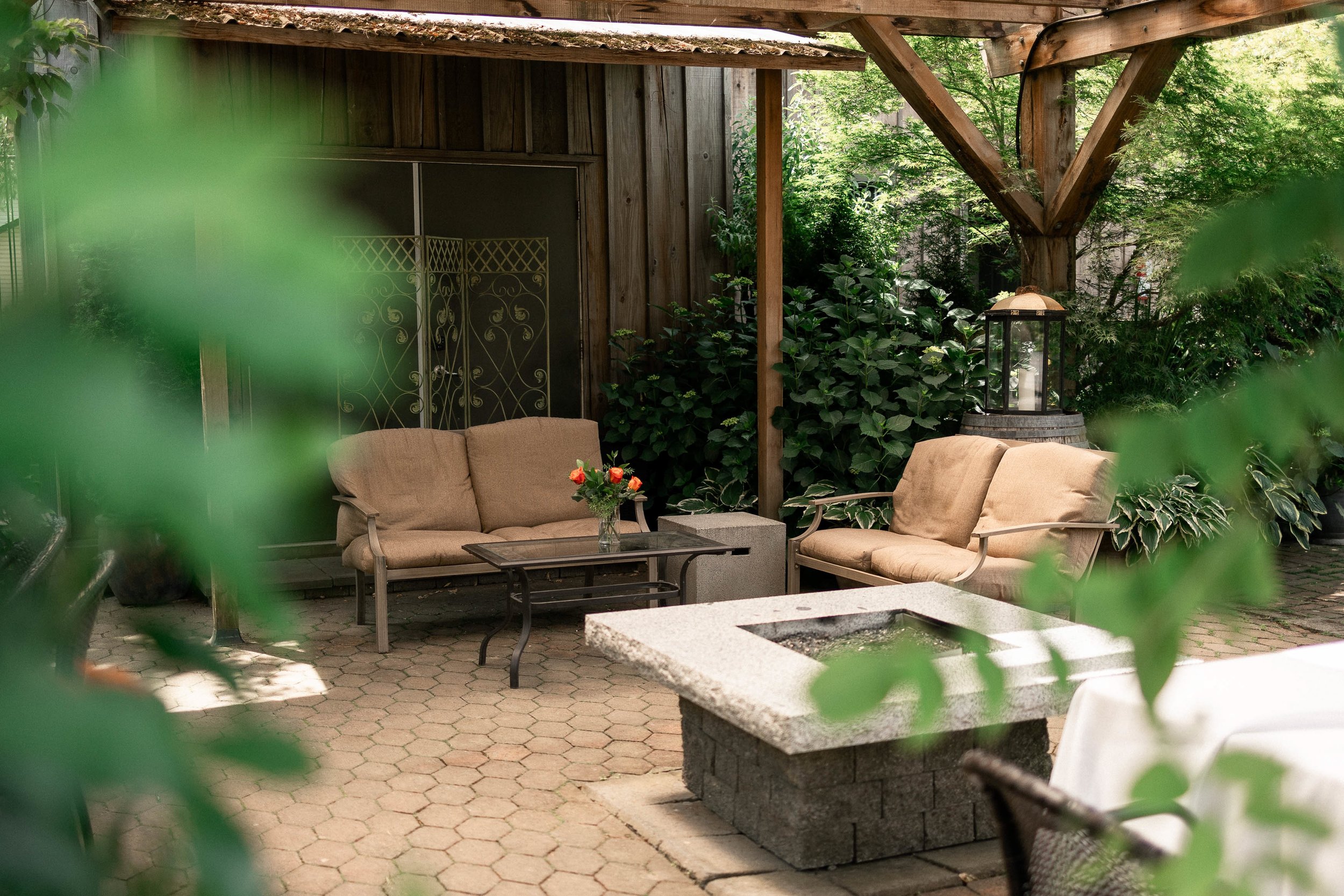 An outdoor patio area with two beige cushioned sofas, a coffee table with a vase of orange and red flowers, surrounded by green foliage. There is a stone fire pit in the foreground and a lantern on a barrel behind the sofas. The area is shaded under a wooden pergola or roof.